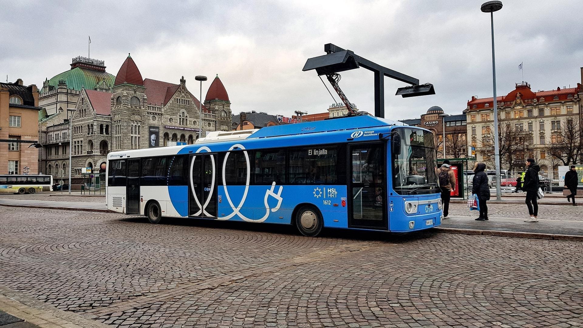 Photograph of a electric bus stopped at the bus station in the middle of a beautiful city with old buildings.