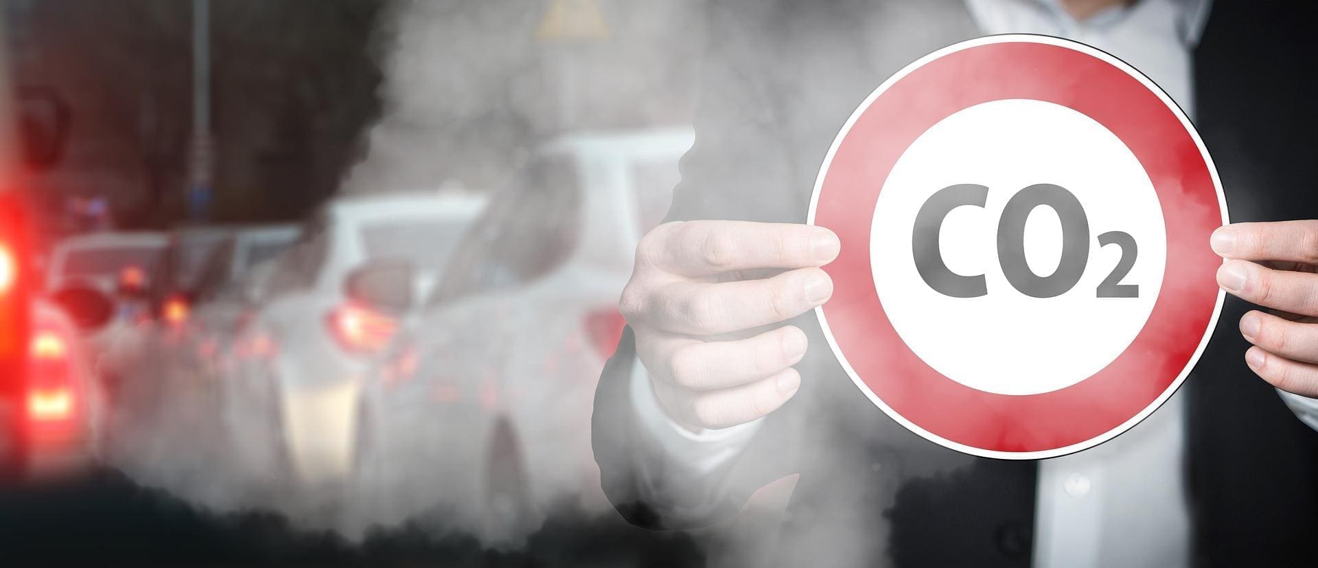 Photograph of a man holding in his hands a limiting speed shield, on which it is written: CO2. In the background are many car, stuck in traffic jam, with their motors running and producing CO2.