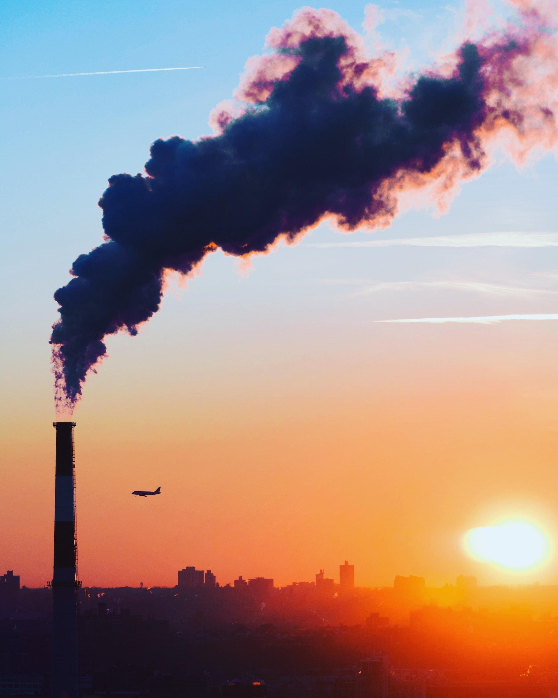 Photograph of a industrial company releasing CO2 into the atmosphere. In the backgroung, on the sky, a plane is flying and the sun is setting.