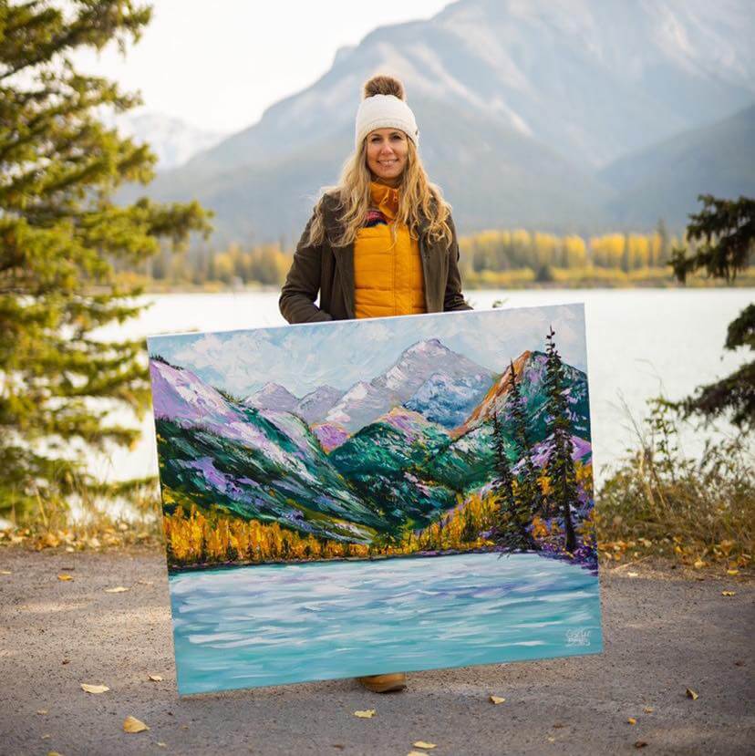 Photograph of Giselle Denis holding one of her paintings. She finds herself in nature, and the landscape in her background is represented in her painting.