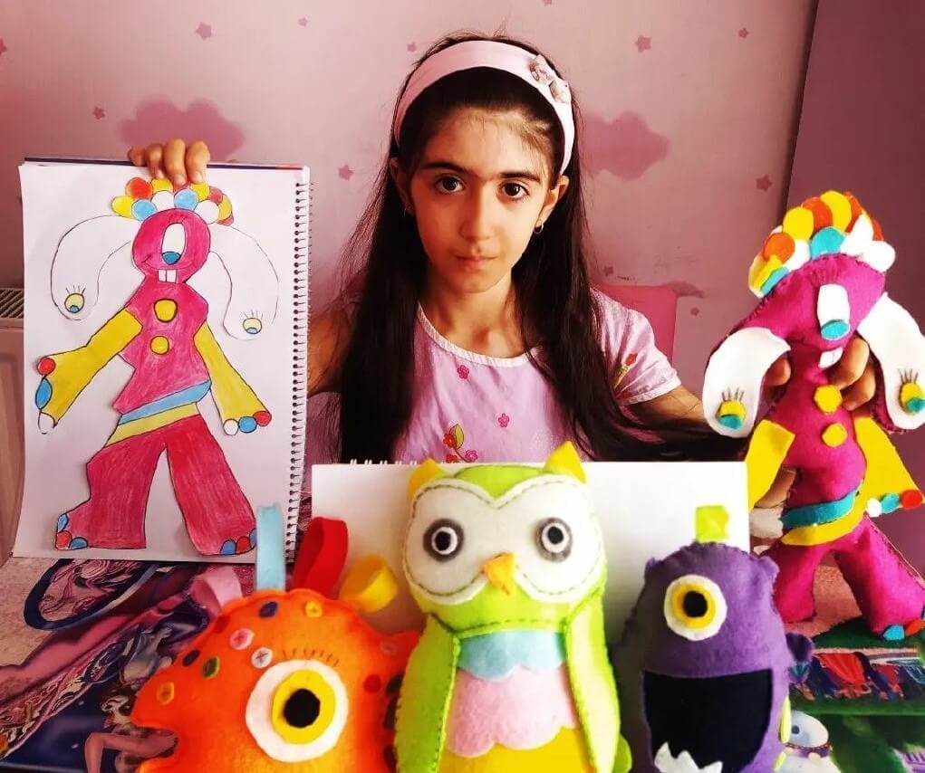 Photograph of Ronia Khalaj in her room, surrounded by the toys she created herself. With one hand, she holds the sketch she used to create one of her toys. On the other hand, she holds the created toy.
